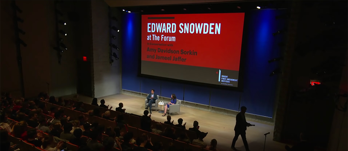 Auditorium with two speakers seated on stage and a slide announcing Edward Snowden at The Forum.